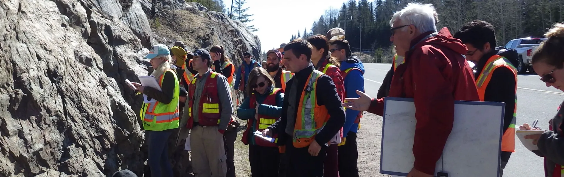 Several students outside by a rock cliff examining the minerals while a prof is observing.