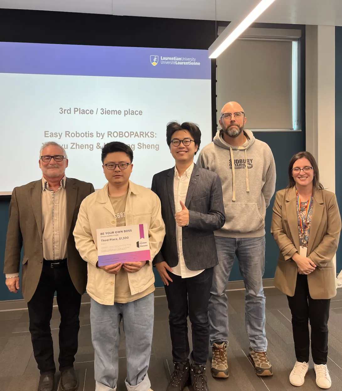 Third-place BYOB winners Xingyu Zheng and Haoliang Sheng of ROBOPARKS stand with their award certificate alongside three Laurentian University representatives. The background screen displays '3rd Place / 3ieme place - Easy Robotics by ROBOPARKS.