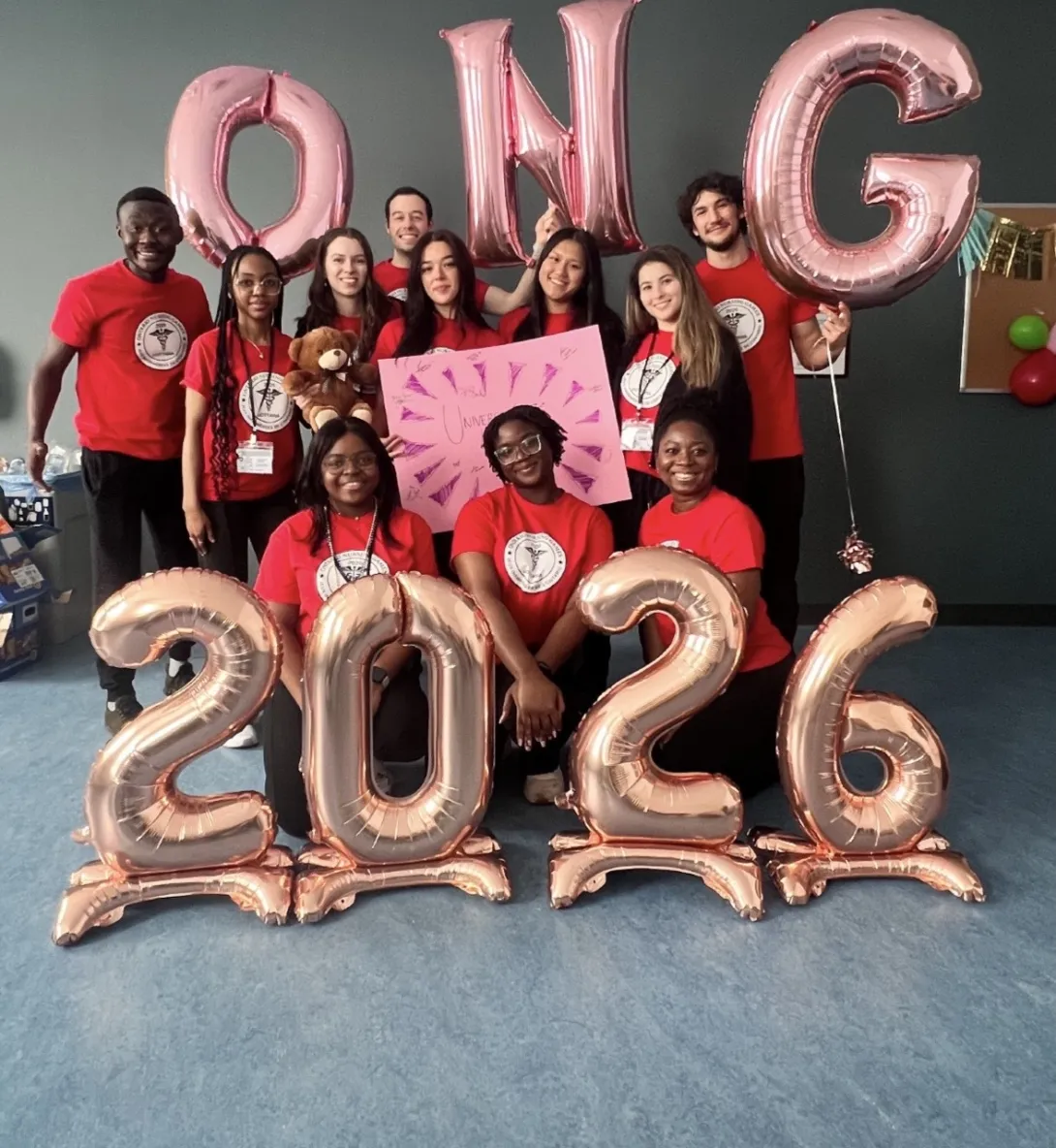 A group of eleven celebratory nursing students from Laurentian University poses indoors with rose-gold foil balloons. The students are dressed in matching red T-shirts featuring a circular medical logo. In the background, three students hold balloons spelling "ONG" (Ontario Nursing Games). In the foreground, four large floor-standing balloons spell out "2026."