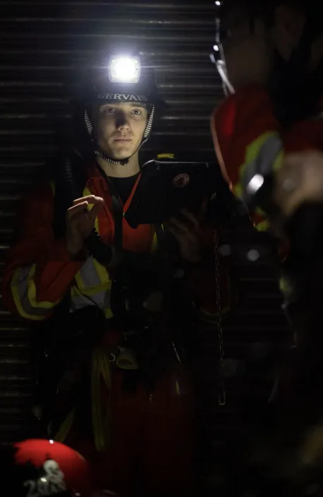 Félix Gervais, President of the Laurentian University Mine Rescue Club, looks at a digital tablet while participating in a mine rescue simulation. He is wearing high-visibility gear and a black hard hat equipped with a glowing headlamp that illuminates the dark environment.