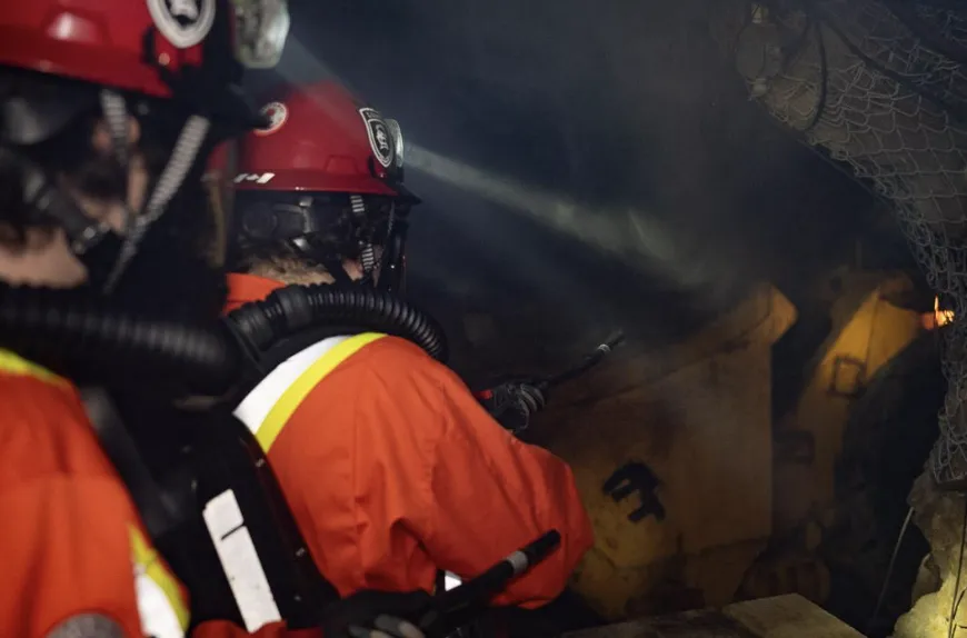 Laurentian University Mine Rescue team members work in a dark, smoky underground environment during a competition simulation. The students are wearing red helmets with headlamps and breathing apparatuses while using handheld tools near a piece of heavy mining machinery.