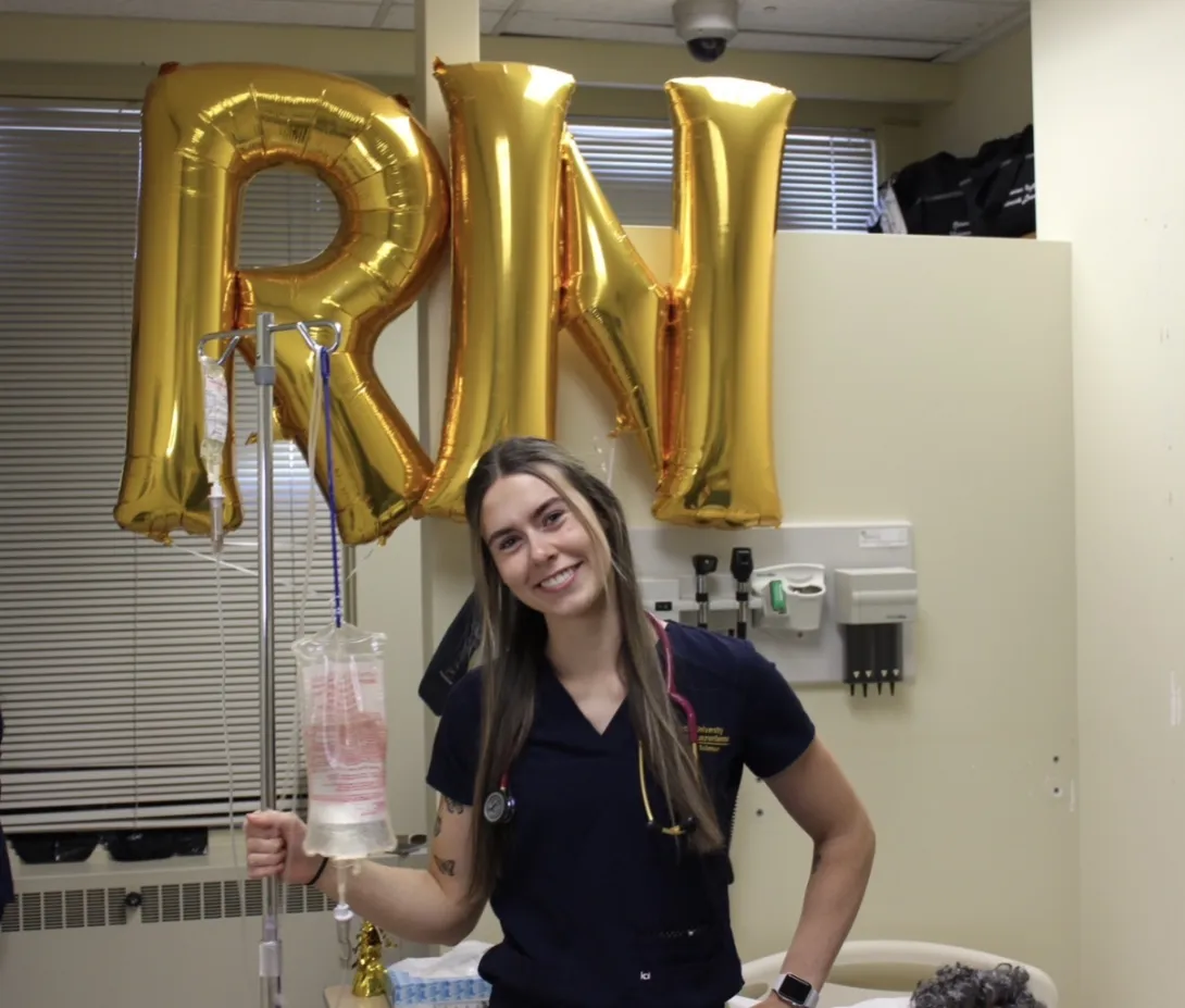 Alex Kowalski celebrating becoming a Registered Nurse. She is smiling in dark navy scrubs with a stethoscope, leaning on an IV pole in front of large, metallic gold foil balloons that spell out "RN".