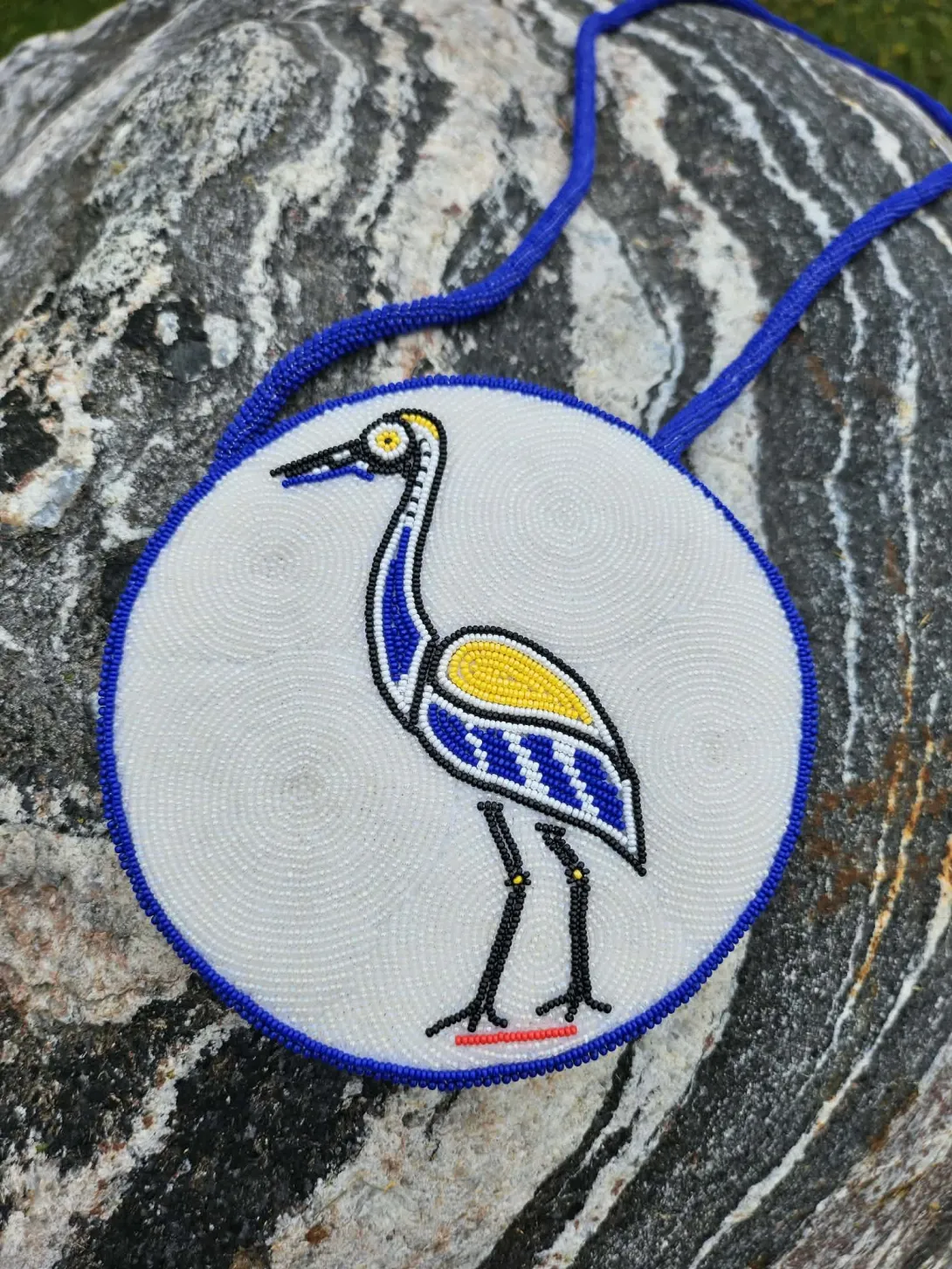 A circular, custom-beaded Chancellor’s medallion resting on a large, textured gray rock. The medallion features a white beaded background with a detailed blue, yellow, black, and white crane in the center, representing the Ajijaak Doodem (Crane Clan). It is framed by a vibrant blue beaded border and attached to a matching blue beaded strap.
