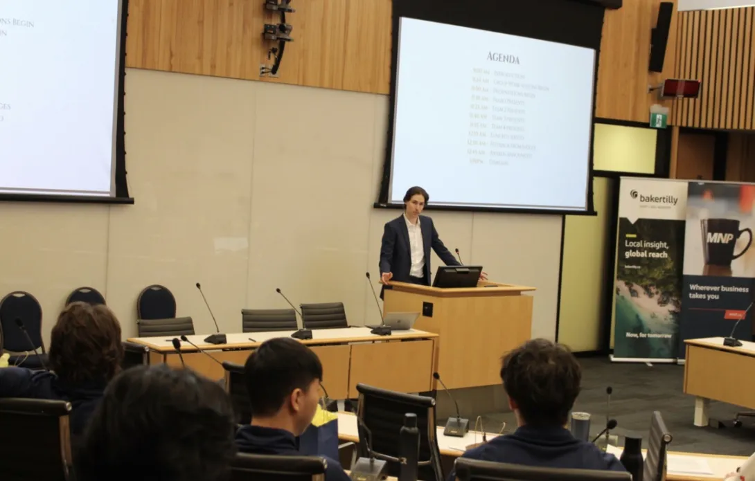 Paul Petitclerc stands at a wooden podium at the front of a large, modern university lecture hall, delivering a presentation. He is wearing a blue suit jacket and open-collared white shirt, gesturing with his hands. Large projection screens behind him display a slide titled "AGENDA," while several students sit at rows of desks in the foreground listening to him speak.