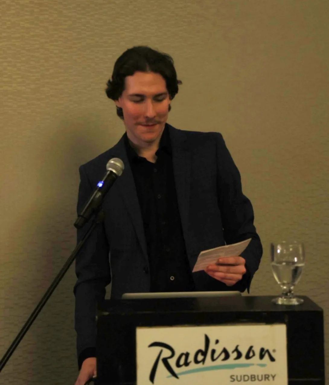 A close-up photograph of Paul Petitclerc speaking at a podium during a formal event. He is wearing a dark navy blue suit blazer over a black button-down shirt. He is looking down slightly at a small note card in his hand with a microphone positioned in front of him. A black sign on the front of the podium reads "Radisson SUDBURY."