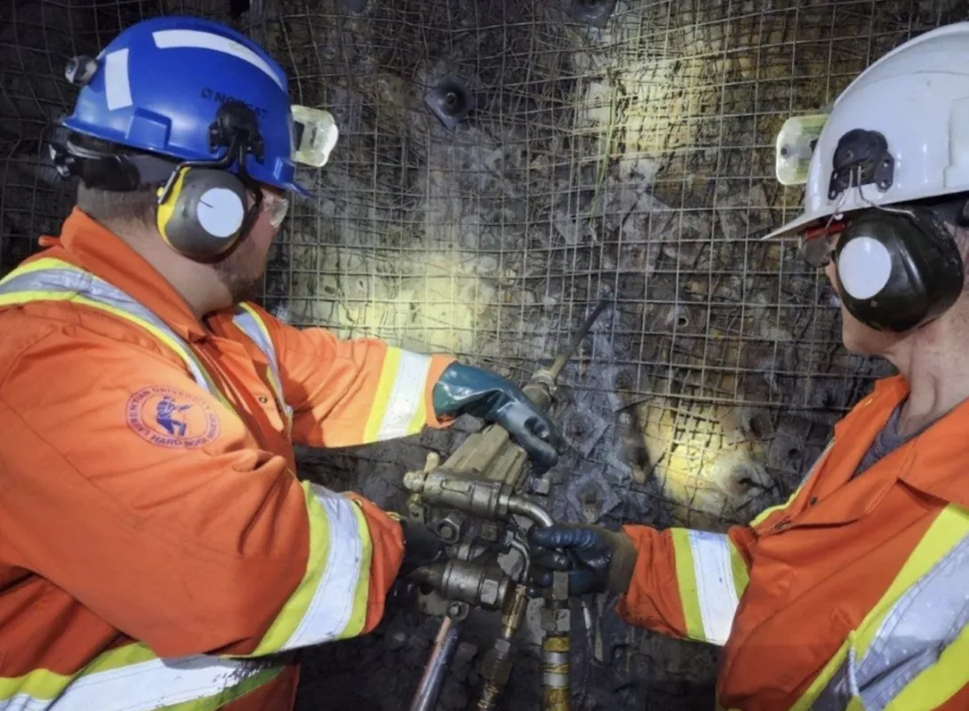 Two miners working underground in a hard-rock mine, wearing high-visibility orange and yellow reflective safety jumpsuits, hard hats, safety glasses, and ear protection. The miner on the left wears a blue hard hat featuring a "Laurentian University Hard Rock Miners" crest on the jacket sleeve, operating a heavy piece of drilling equipment against a rock wall secured by wire mesh. The miner on the right wears a white hard hat and assists in stabilizing the equipment.