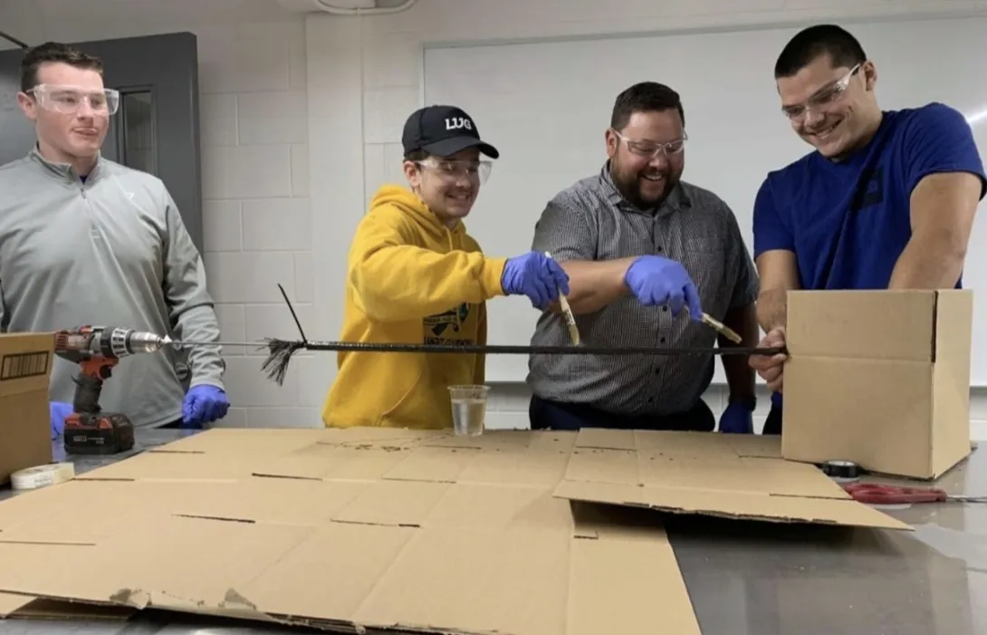 Four male engineering students collaborating in a laboratory setting around a stainless steel table protected by cardboard sheets. In the centre, two students wearing safety glasses and blue disposable gloves use brushes to apply a coating to a long, black structural rod. 