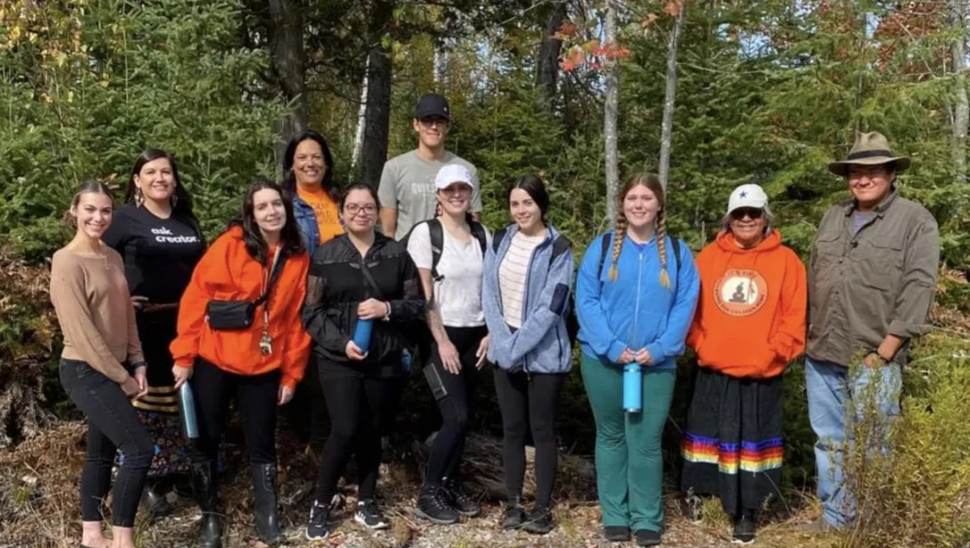 A group of eleven people standing together outdoors on a gravel path with a lush forest backdrop. Kailey O'Bumsawin stands third from the left wearing a bright orange hoodie and black pants. 
