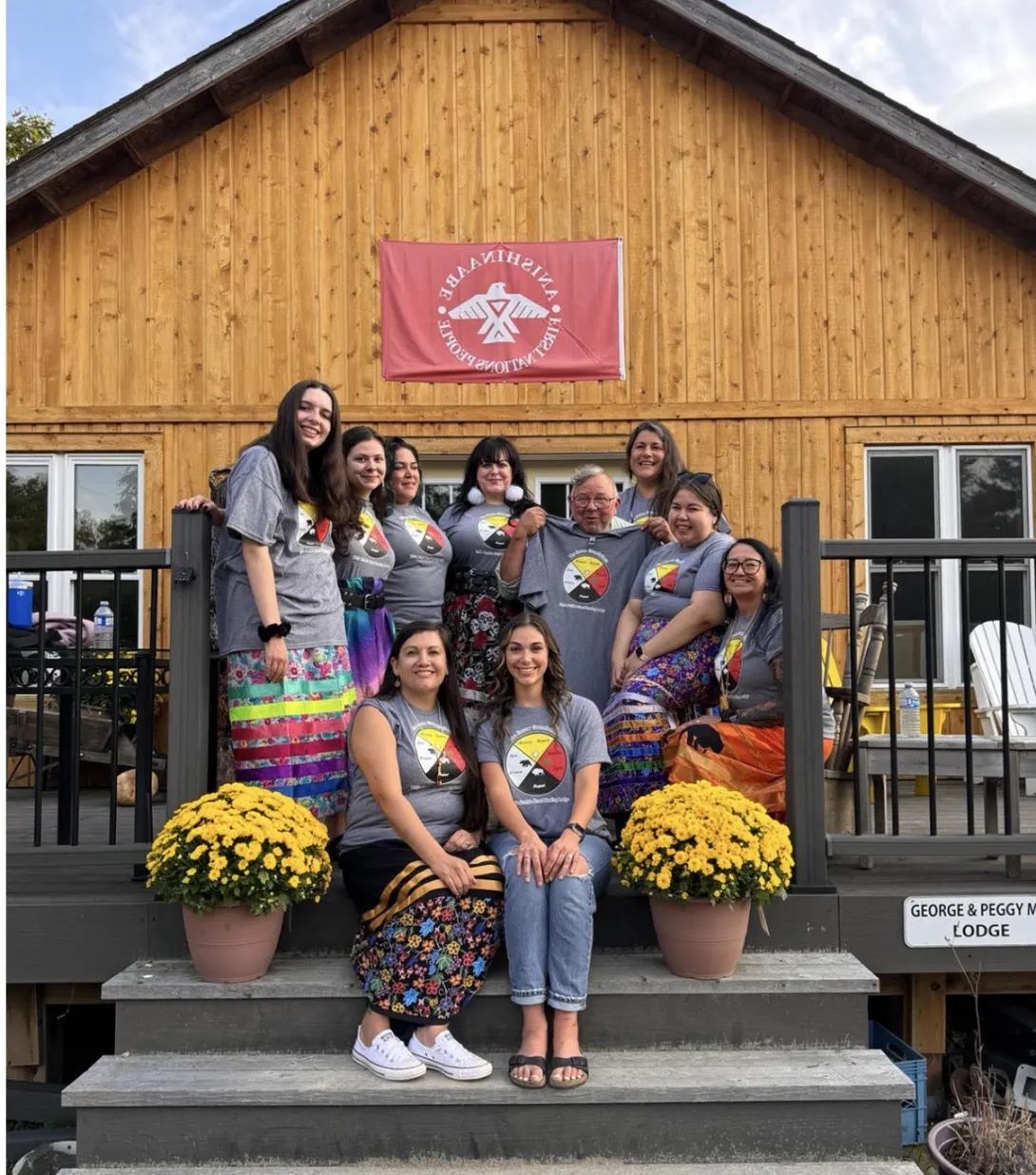 A group of ten people posing together on the wooden steps and porch of the "George & Peggy Manuel Lodge." A red flag reading "Anishinabe First Nations People" with a white thunderbird logo hangs on the cabin wall above them. Most group members are wearing matching grey t-shirts with a traditional four-color medicine wheel graphic, paired with colorful, patterned ribbon skirts.