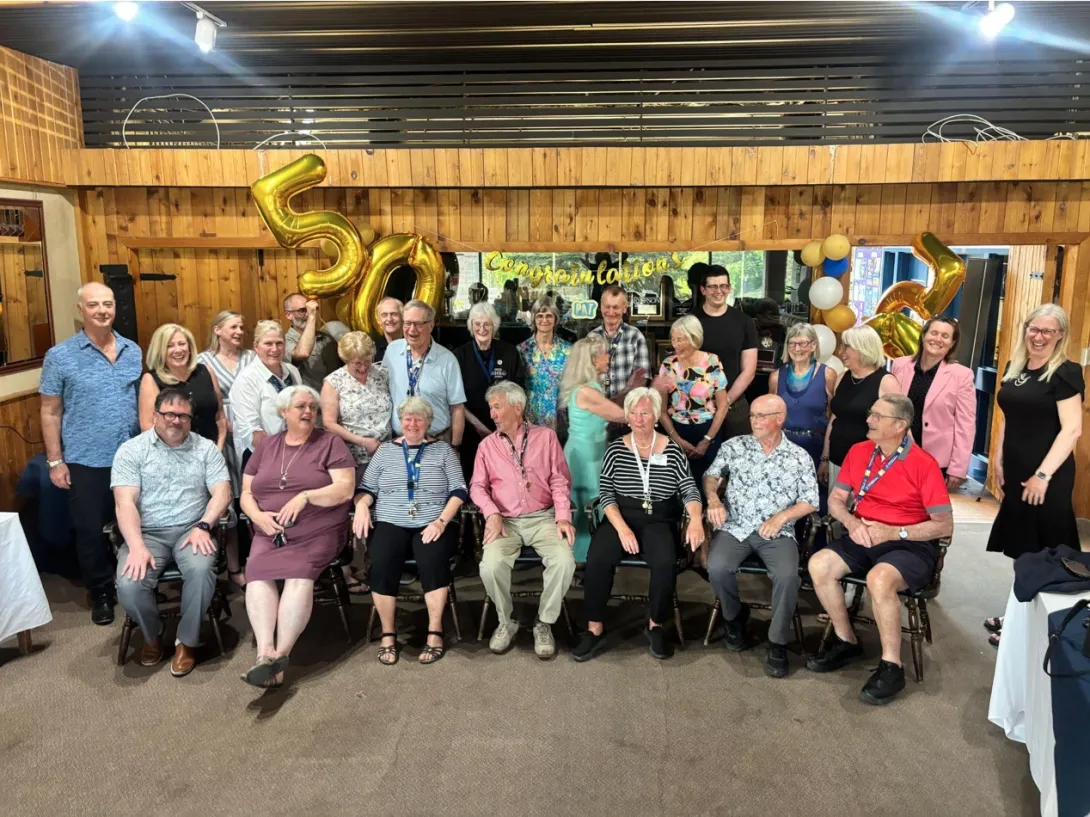 A large, cheerful group of alumni and friends pose for a photo in the Ken Bahnuk lounge. Gold foil balloons forming the number "50" and a banner reading "Congratulations" hang in the background.