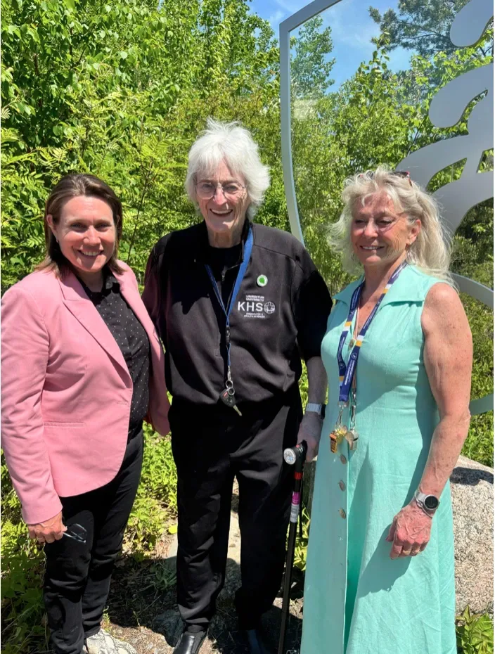 Dr. Pat Pickard stands smiling in the center wearing a black Laurentian University Kinesiology jacket. She is flanked by Sarah Stevens on her right in a pink blazer, and Erica Carlson on her left wearing a mint-green dress and athletic whistles. Both of these individuals received the exceptional alumni award. They are posing outdoors in front of a metallic university crest.