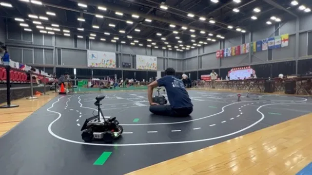A wide view of an indoor gymnasium arena where a small autonomous vehicle drives along a black track marked with white lane lines. A team member sits on the floor near the track monitoring the robot, with international flags hanging in the background.