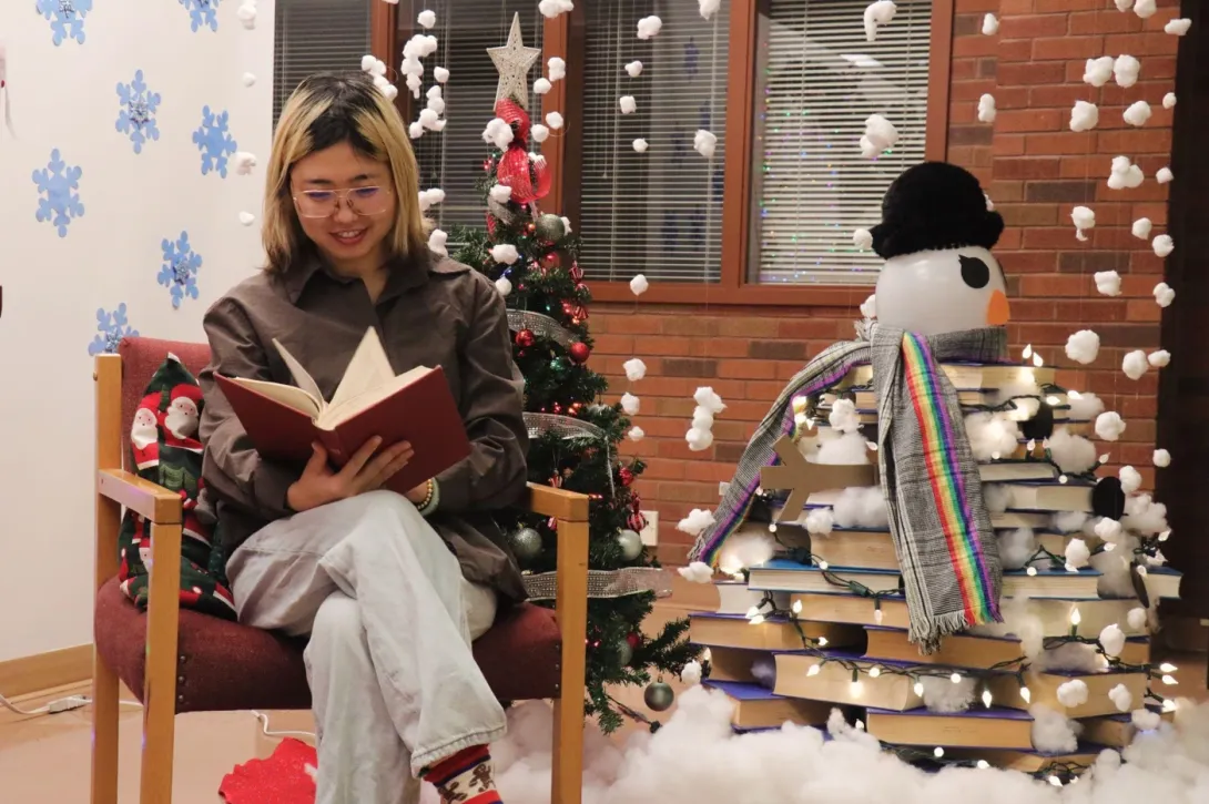 Felice Guo sitting in an armchair inside the J.N. Desmarais Library at Laurentian University, smiling while reading a book. The space is decorated for the holidays with a small Christmas tree and a creative snowman made entirely out of stacked library books wearing a black hat and a rainbow scarf.