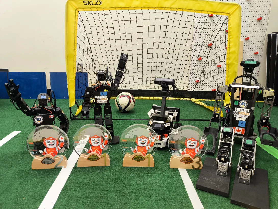 Four FIRA RoboWorld Cup awards—two 1st Place, one 2nd Place, and one 3rd Place—displayed on a green turf field in front of a small soccer net. Four humanlike and autonomous SnoBots robots stand behind the trophies, positioned around a soccer ball.