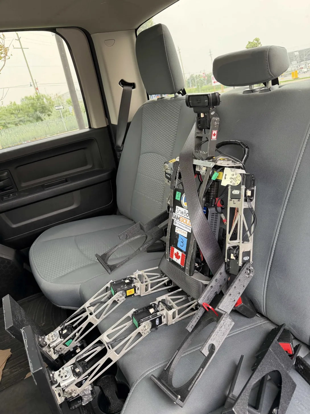 A tall, silver-and-black humanoid robot sitting securely in the back seat of a truck, buckled in with a standard seatbelt. The robot features sponsor decals including IAMGOLD and KGHM, along with a Canadian flag sticker.