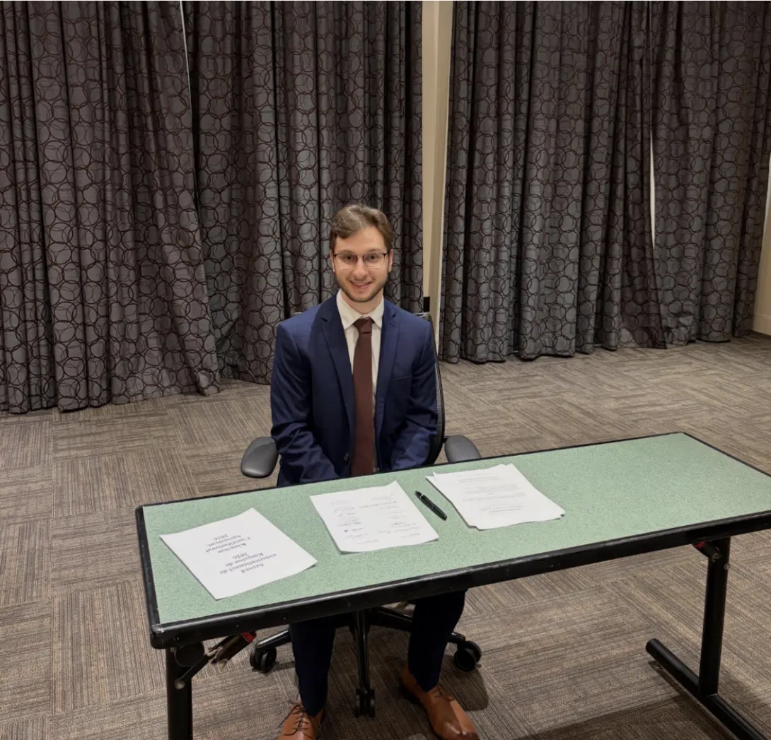 Matteo Rocca sits smiling at a green conference table in front of patterned curtains. He is wearing a navy blue suit with a dark red tie. Spread across the table are pages from the finalized 2026 Kingston Constitutional Agreement.