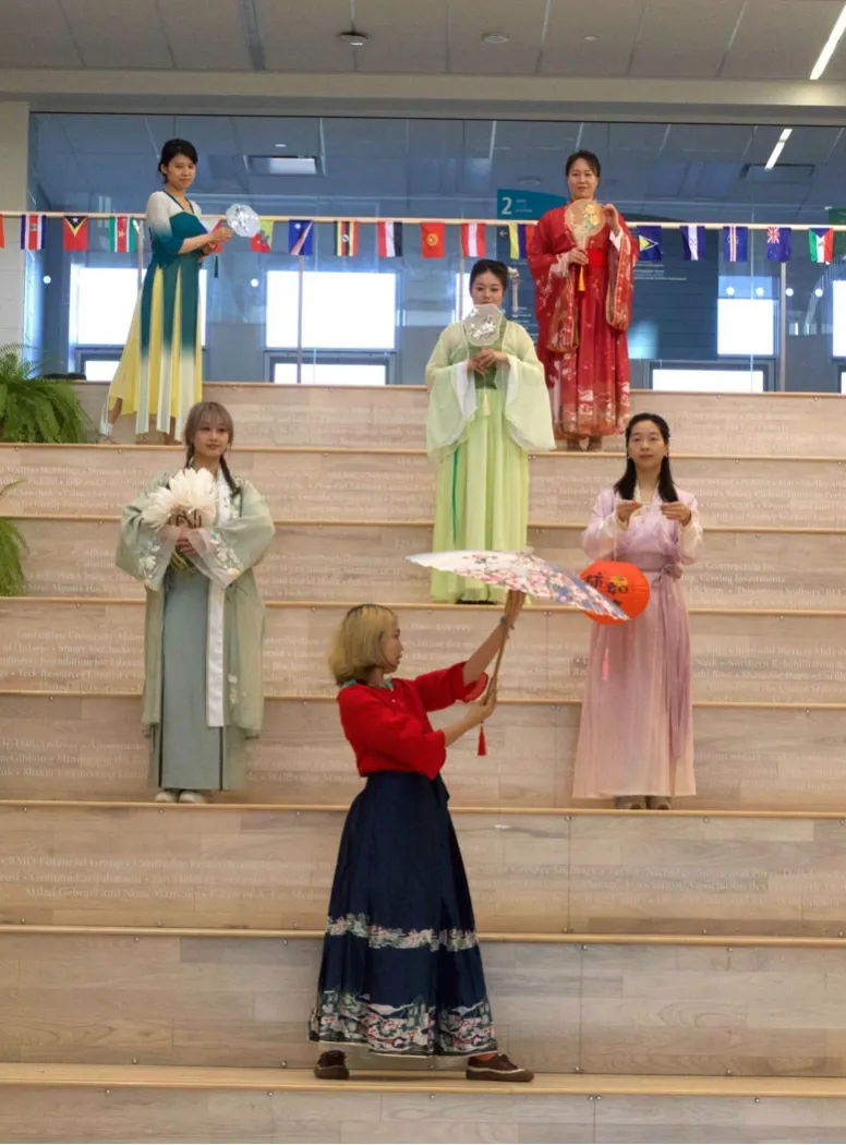 Six women posing on the large wooden steps of the Parker Atrium at Laurentian University, showcasing traditional Chinese Hanfu dresses in vibrant shades of red, green, pink, and yellow. They are holding traditional fans and a paper umbrella, with an array of international flags hanging from the balcony railing above them.