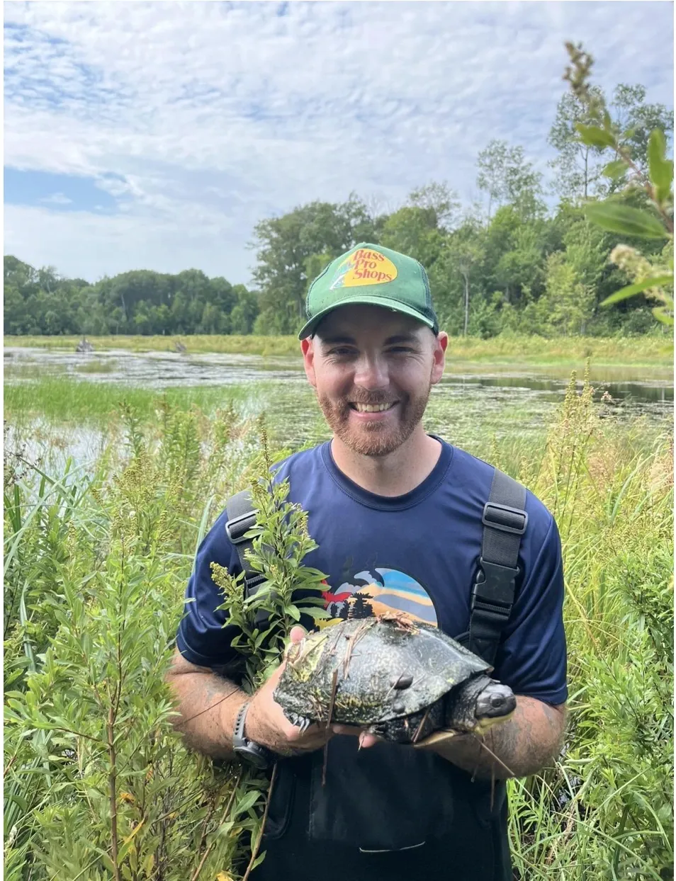 A close-up portrait of Aidan Maloney smiling in a green cap and chest waders, holding up a large Blanding's Turtle covered in wetland debris, with a lush marsh and blue sky in the background.