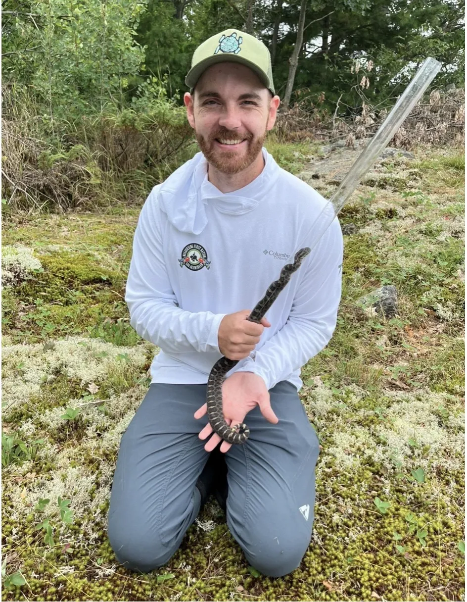 Rear view of biologist Aidan Maloney wearing high-visibility orange safety gear, conducting radio-telemetry to locate a tracked turtle over a serene wetland lake under a clear blue sky.