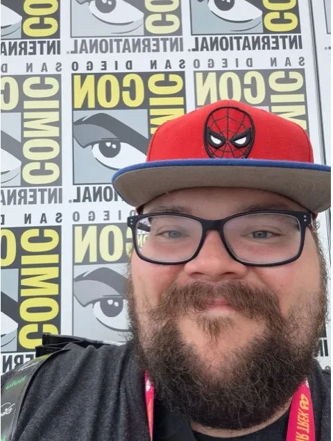 A close-up selfie of Chris Hogge, a man with a full beard and mustache, wearing black-framed eyeglasses and a red baseball cap featuring a Spider-Man emblem on the front. He is smiling at the camera outdoors, standing in front of a San Diego Comic-Con backdrop patterned with yellow and black text and eye logos.