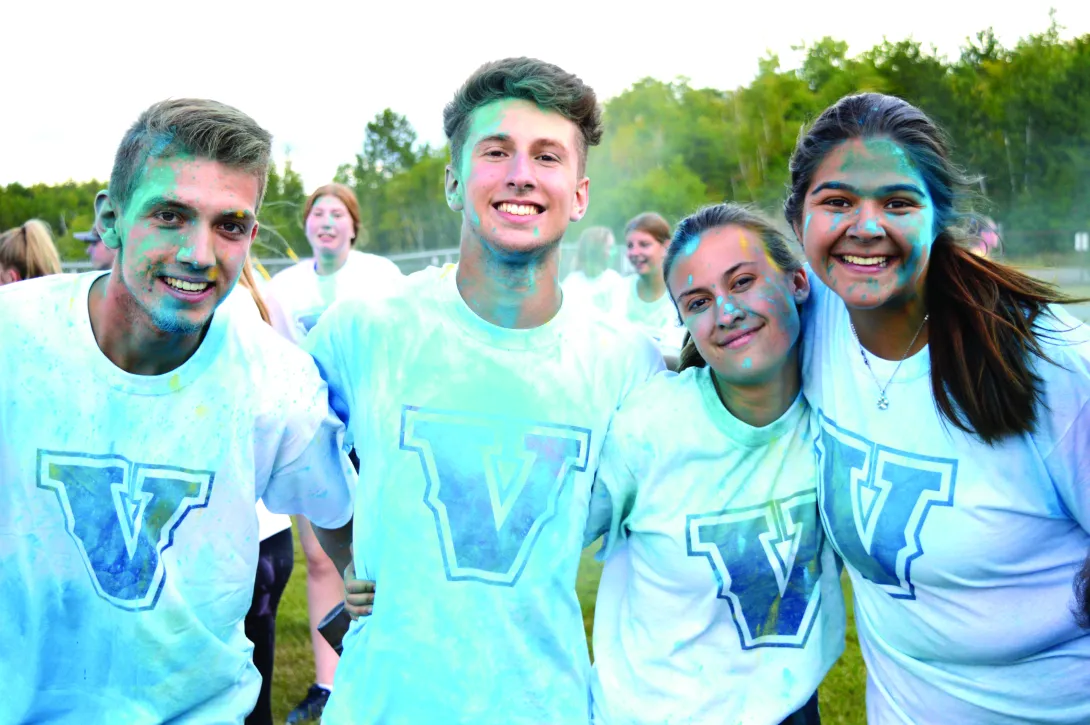 Four smiling Laurentian University students posing together outdoors during an orientation color run event. They are wearing matching light blue t-shirts featuring a large, stylized blue "V" logo, with blue and yellow powder paint splattered across their shirts, faces, and hair.