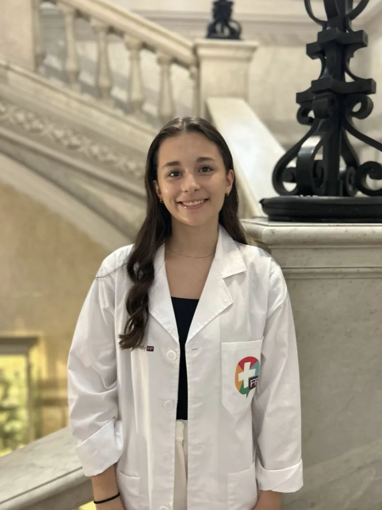 Karina Schryer smiling while wearing a white medical lab coat over a dark shirt, posing in front of an ornate marble staircase and wrought-iron railing.