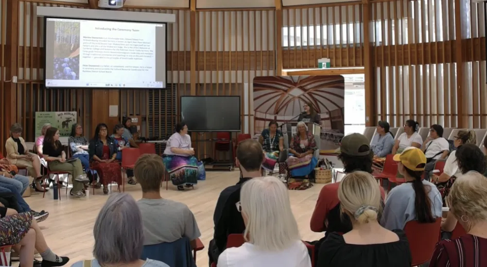 Community members and local leaders gather in a circle inside the Indigenous Sharing and Learning Centre at Laurentian University during the project launch and feast event in June, with a project facilitator presenting at a podium in the background.