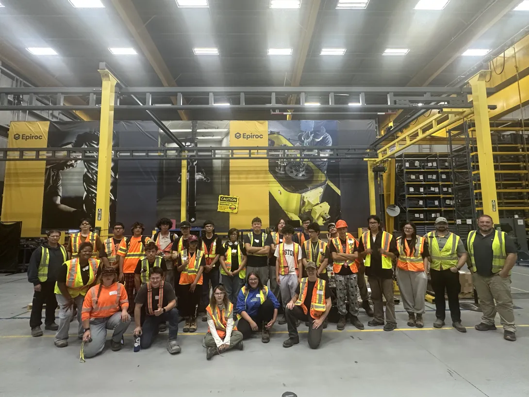 A large group of OYEP participants wearing safety vests and hard hats posing together inside the Epiroc Canada industrial facility.