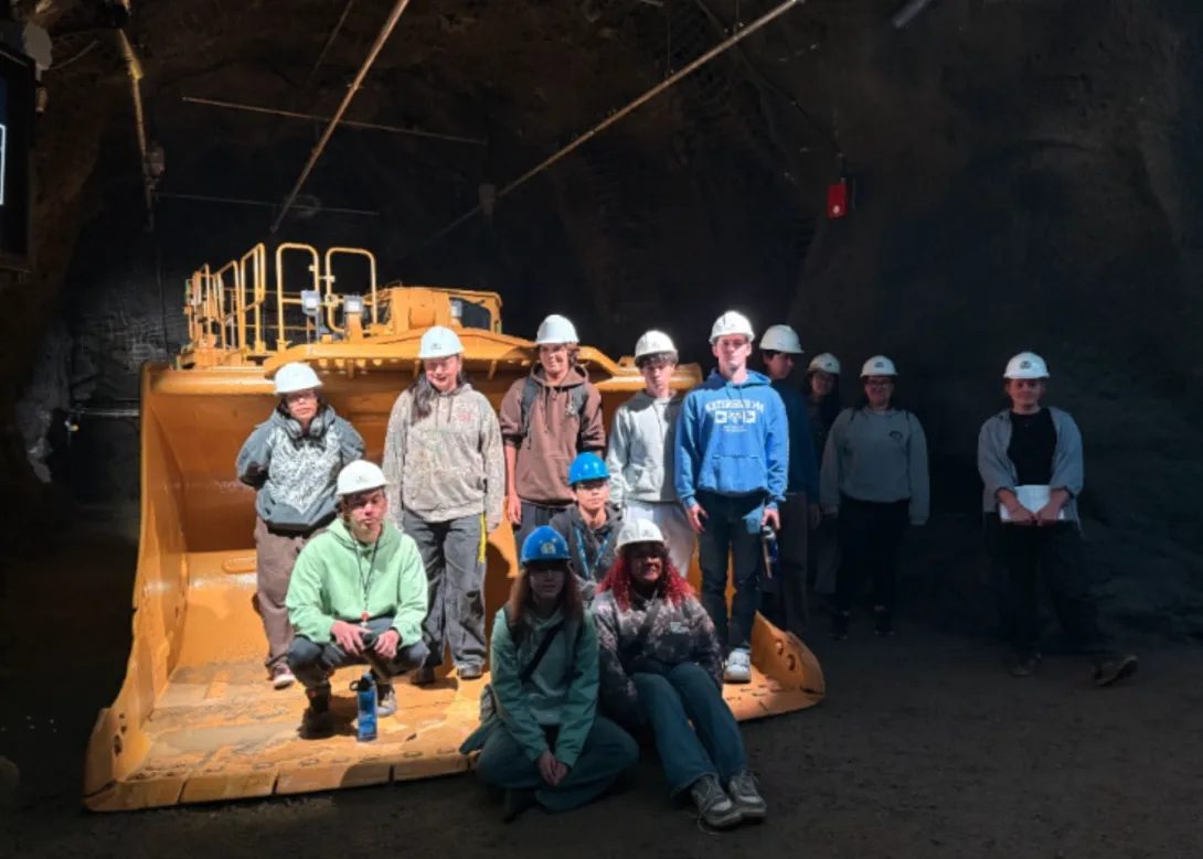 OYEP Rangers wearing hard hats posing in front of a giant yellow underground mining loader inside a dark underground mine tunnel. 