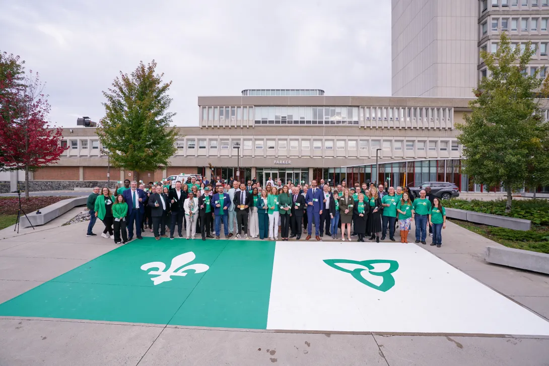 A large group of people wearing green gathering on campus behind a giant Franco-Ontarian flag painted on the courtyard pavement.