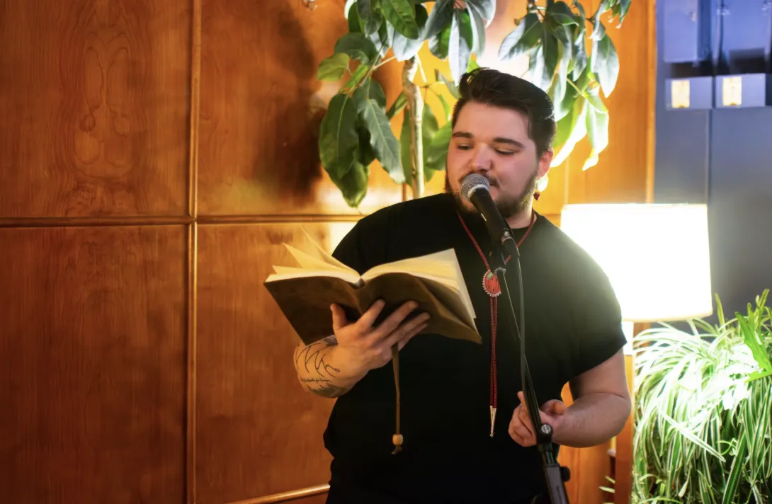 Connor Lafortune reading spoken-word poetry from a leather-bound book into a microphone, wearing a black shirt and a beaded medallion bolo tie.