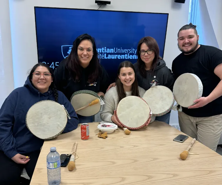 Connor Lafortune seated with his masters cohort around a table at Laurentian University, each holding traditional hand drums.