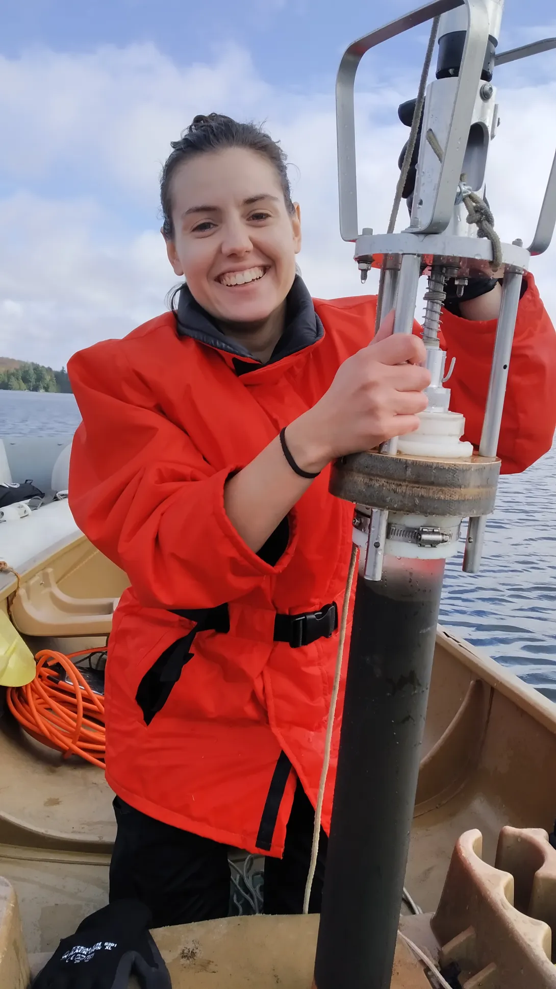 A smiling researcher in a bright red safety coat seated in a boat, holding a metal sediment core sampling device loaded with dark lake mud.