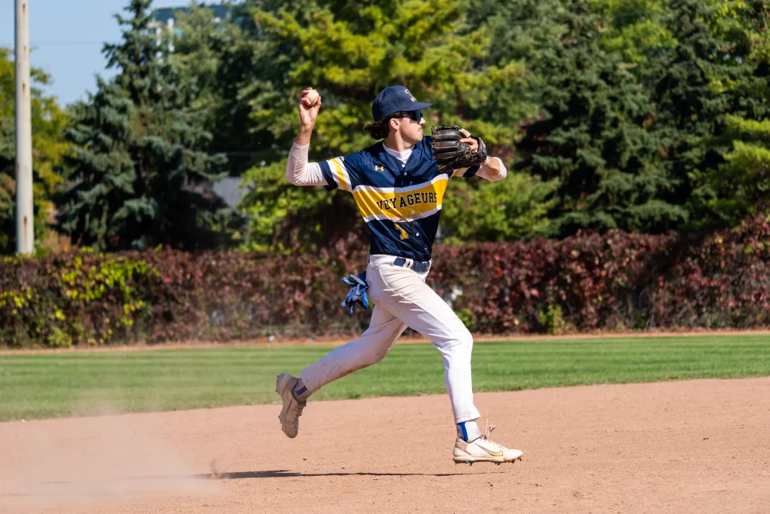 Laurentian Voyageurs shortstop Devan Madore leaping off the dirt field in mid-air to make a throw during a game, wearing a navy and yellow Voyageurs uniform, sunglasses, and a cap.