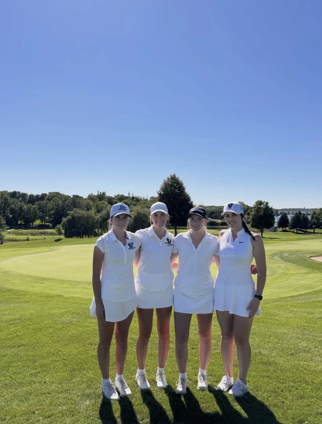 Four female members of the Laurentian Voyageurs golf team stand side-by-side with arms around each other on a sunny golf course under a clear blue sky. All four are dressed in matching white athletic tops, white golf skirts, white caps or visors, and white sneakers, with a green putting area and trees in the background.