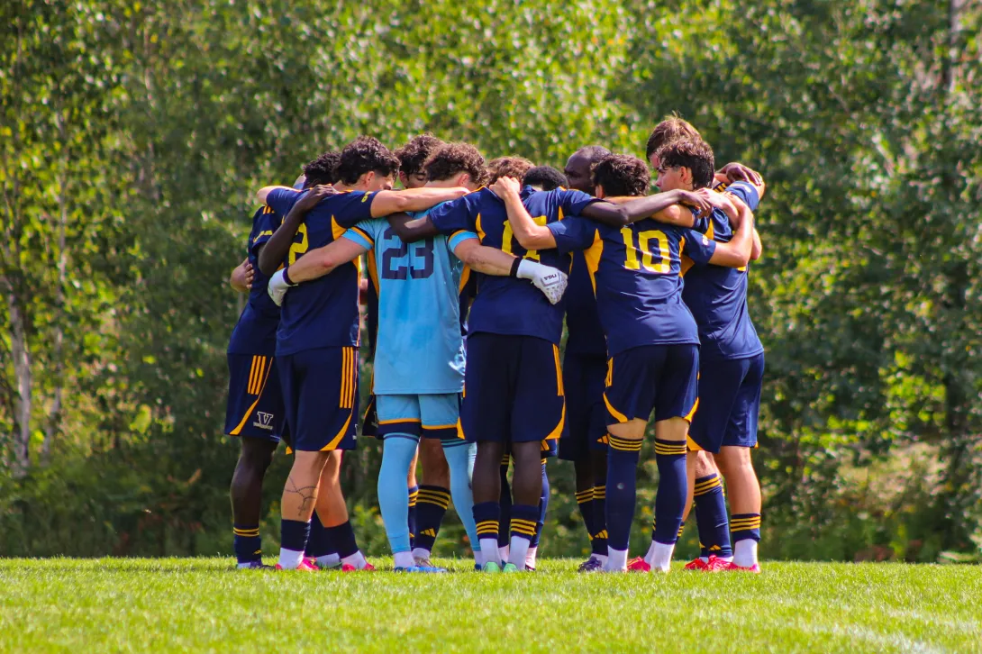 The Laurentian Voyageurs men's soccer team standing in a tight circle on the grass field, arms wrapped around each other's shoulders in a pre-game or post-game huddle, facing away from the camera in their navy and yellow kits alongside their goalkeeper in light blue.