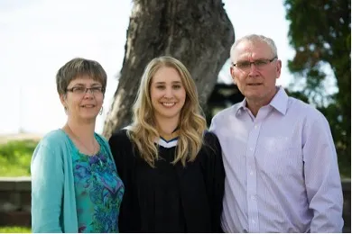Elizabeth Urso smiling in her Laurentian University graduation gown, standing outdoors between her mother and father during convocation.  