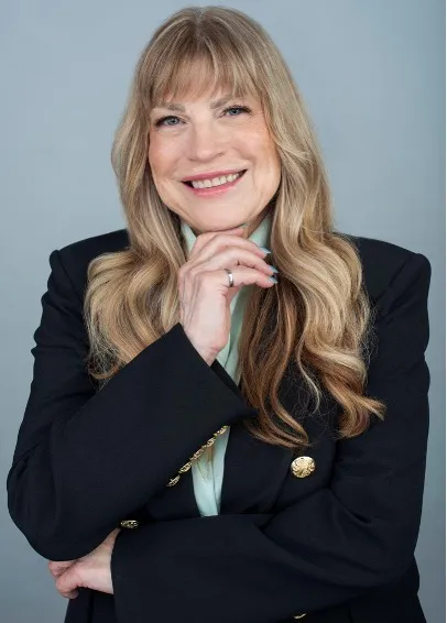 Close-up studio portrait of Rachel Prudhomme smiling with her hand resting gently under her chin, wearing a black blazer with ornate gold buttons, a light green collared shirt, and a silver ring against a plain grey background. 