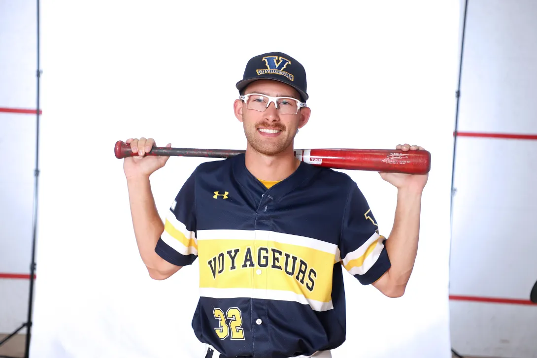 Laurentian Voyageurs senior Ryan Lacasse smiling for a portrait in uniform with glasses, holding a baseball bat behind his shoulders against a white studio backdrop.