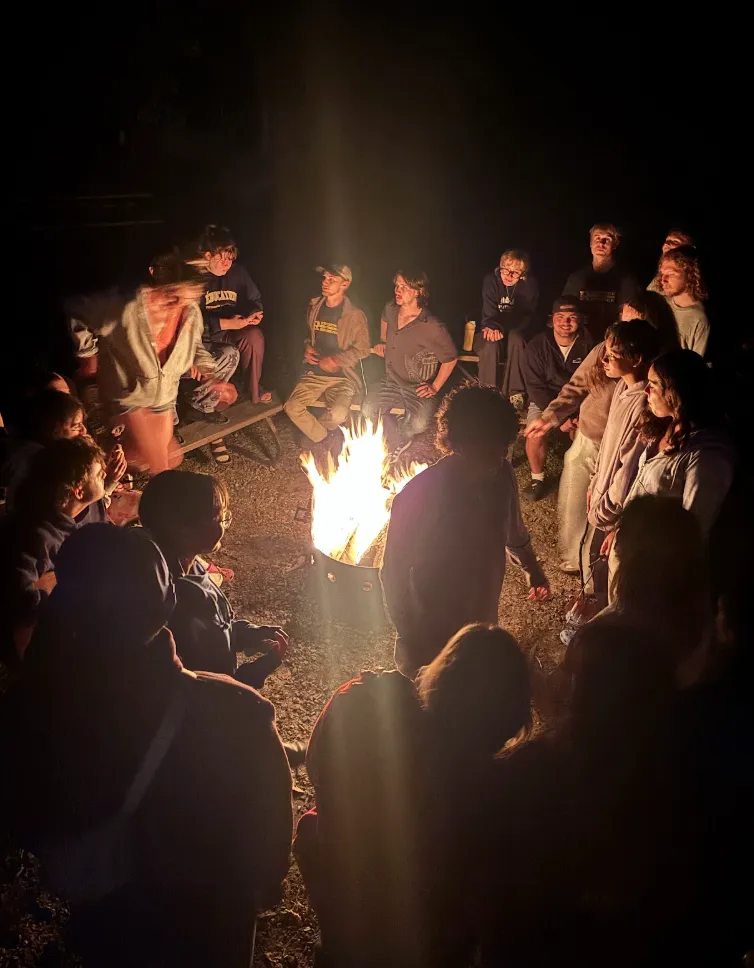 A group of students gathered outdoors at night, sitting on benches and standing in a circle around a glowing campfire
