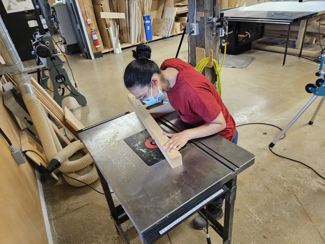 Michelle Zephyr guides a wooden board through a router table in the  McEwen School of Architecture workshop.